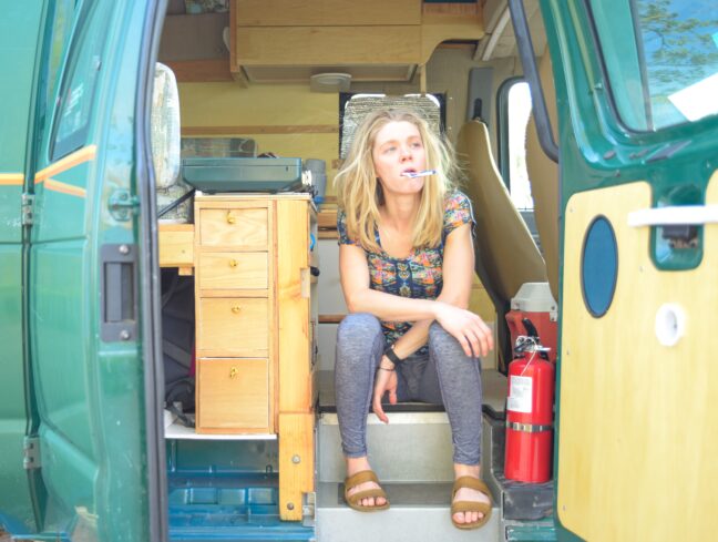 A blonde woman sits in a converter van with the side doors open. She has a toothbrush in her mouth.